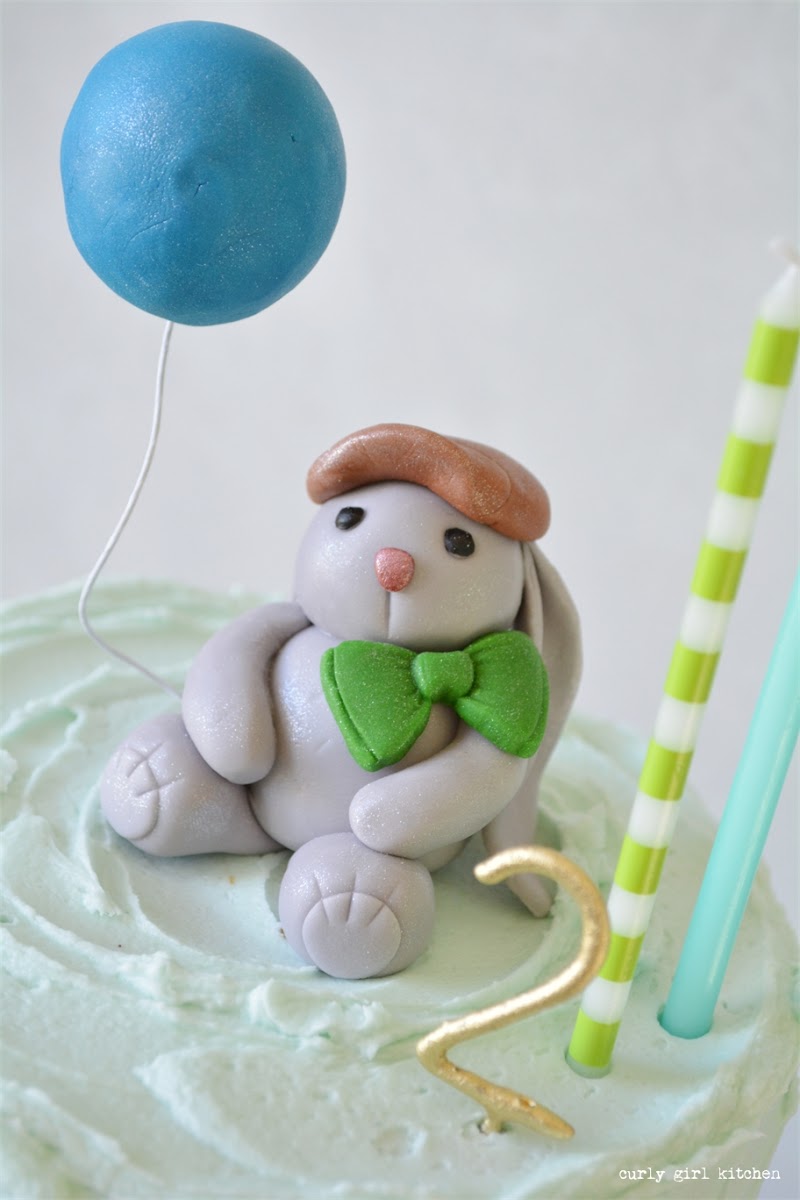 A fondant bunny with a bow and balloon on top of a birthday cake.