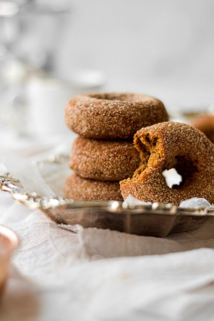 Pumpkin donuts, coated in cinnamon sugar, one with a bite taken out of it, stacked on a silver plate.