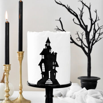 A haunted house layer cake on a black cake stand, with gold candlesticks, black candles and a black tree.