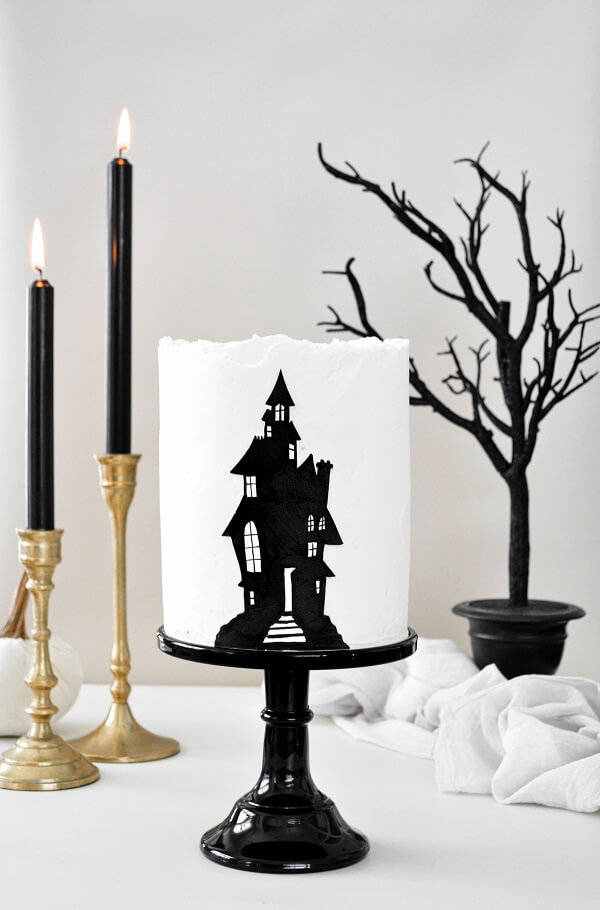 A haunted house layer cake on a black cake stand, with gold candlesticks, black candles and a black tree.