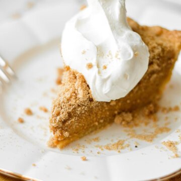 A slice of pumpkin bourbon crumble pie on a white plate, topped with whipped cream, with a yellow linen napkin tucked under the plate.