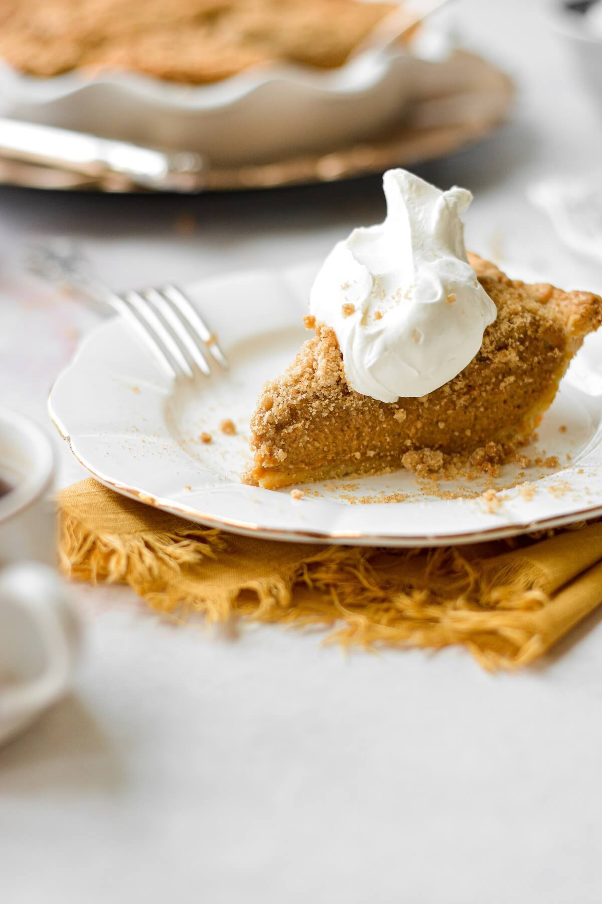 A slice of pumpkin bourbon crumble pie on a white plate, topped with whipped cream, with a yellow linen napkin tucked under the plate.