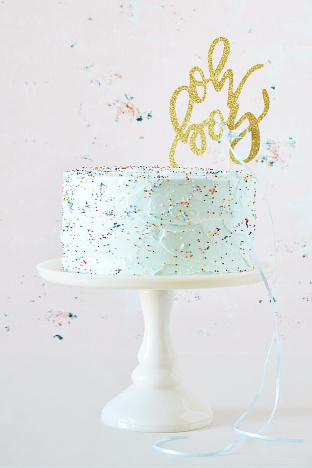 Funfetti Cake with pale blue buttercream, rainbow sprinkles, and a gold "oh boy" cake topper, sitting on a white cake stand.