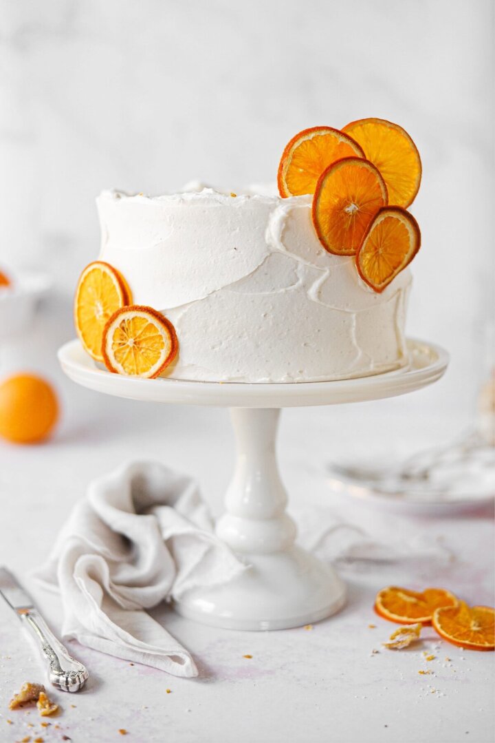 An orange cake, garnished with dried orange slices, on a white cake stand.