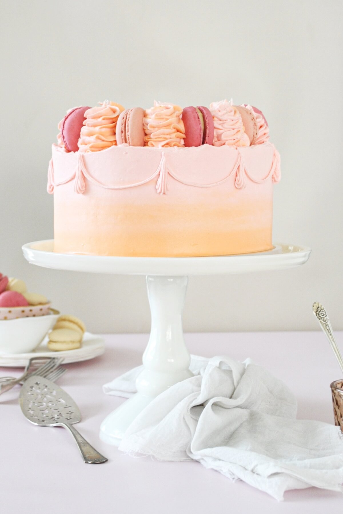 A cake on a white cake stand, frosted with pink and orange watercolor buttercream, topped with macarons and swirls of buttercream.