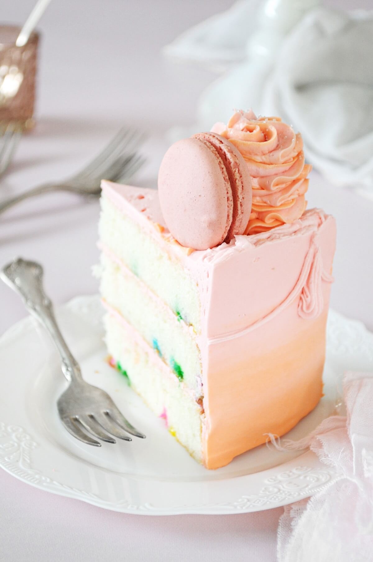 A slice of funfetti cake, frosted with pink and orange watercolor buttercream, topped with a macaron and swirl of buttercream.