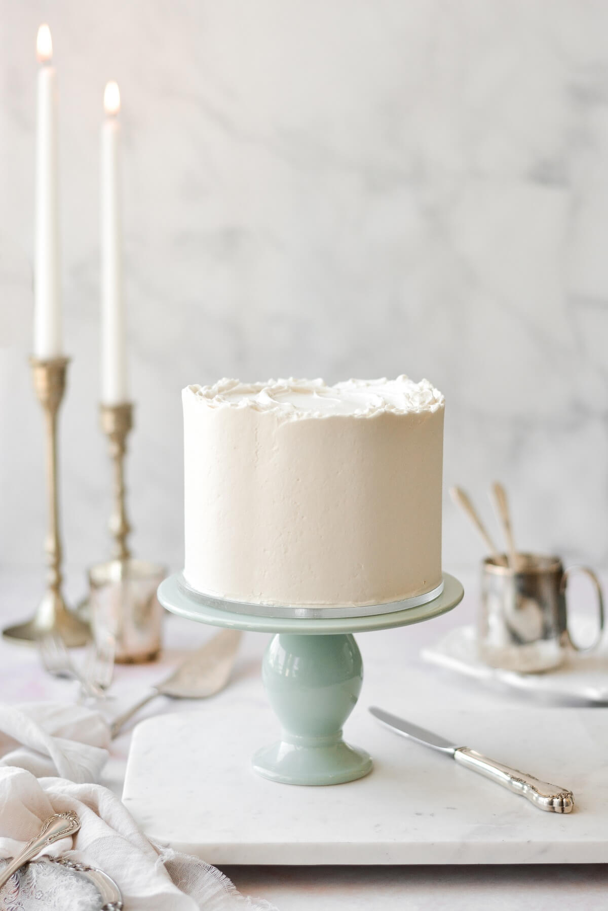 White velvet cake on a sage green cake stand, with taper candles in the background.