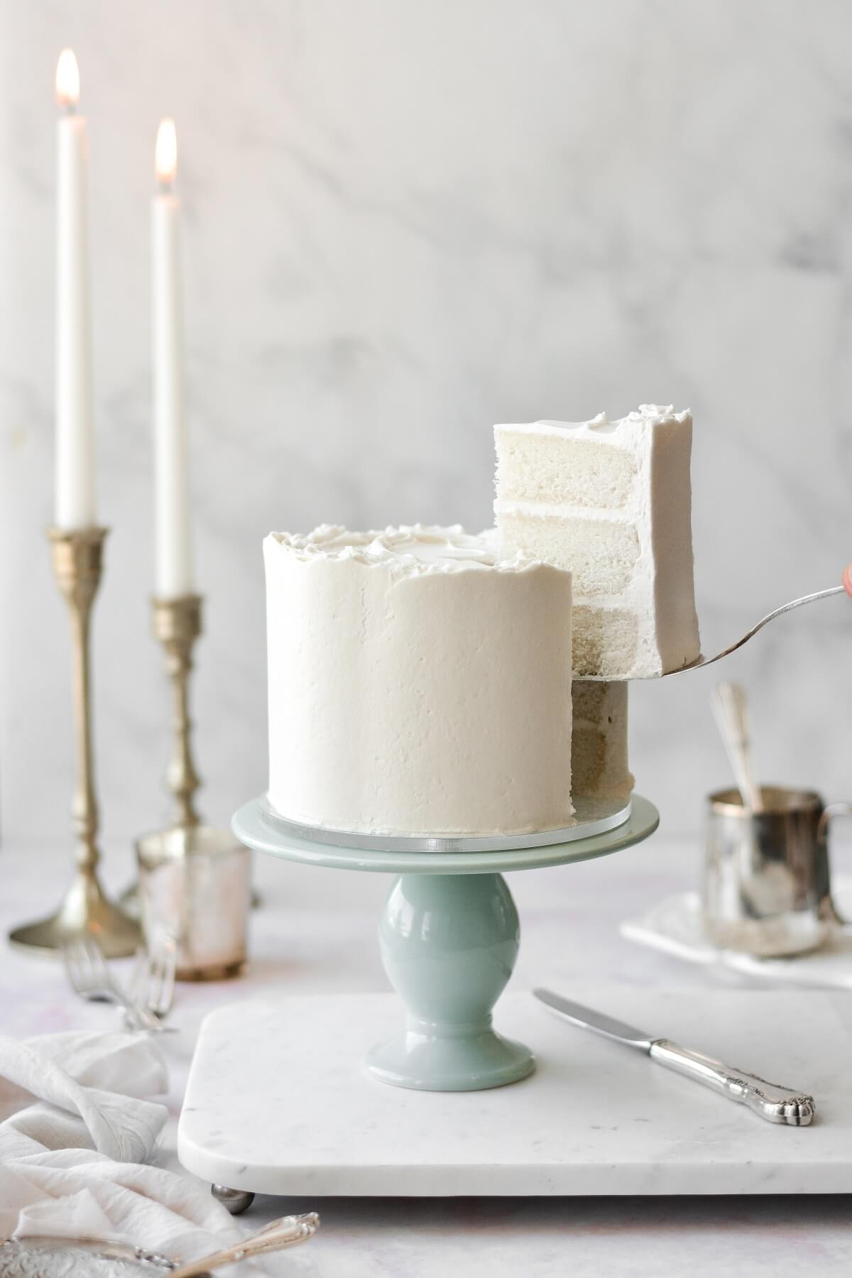 White velvet cake, on a sage green cake stand, with a piece being lifted out by a silver cake server.