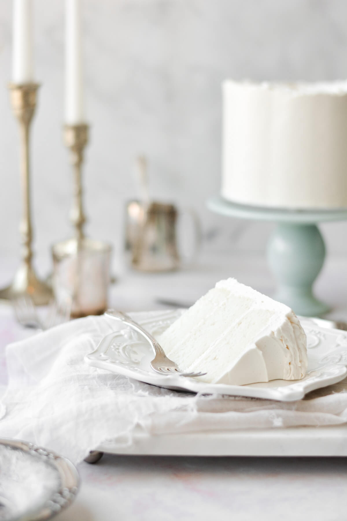 A slice of white velvet cake on a white plate, with a silver fork., and the cake and candles in the background.