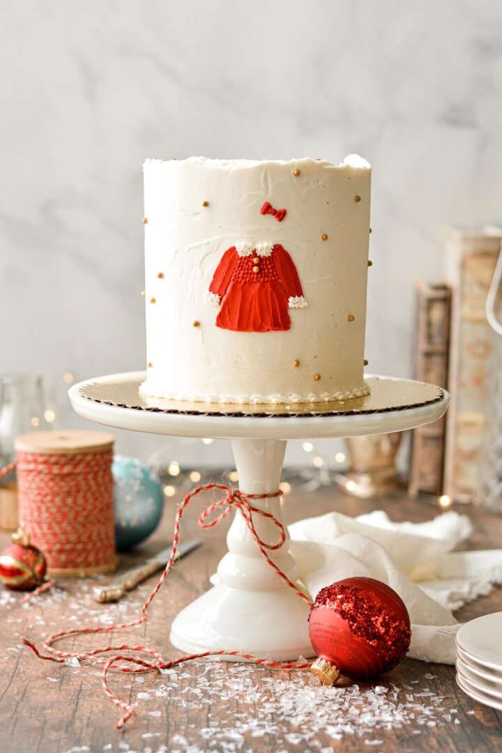 A Christmas cake with gold and red sugar pearls, ornaments, and a red Christmas dress painted on the cake in buttercream.
