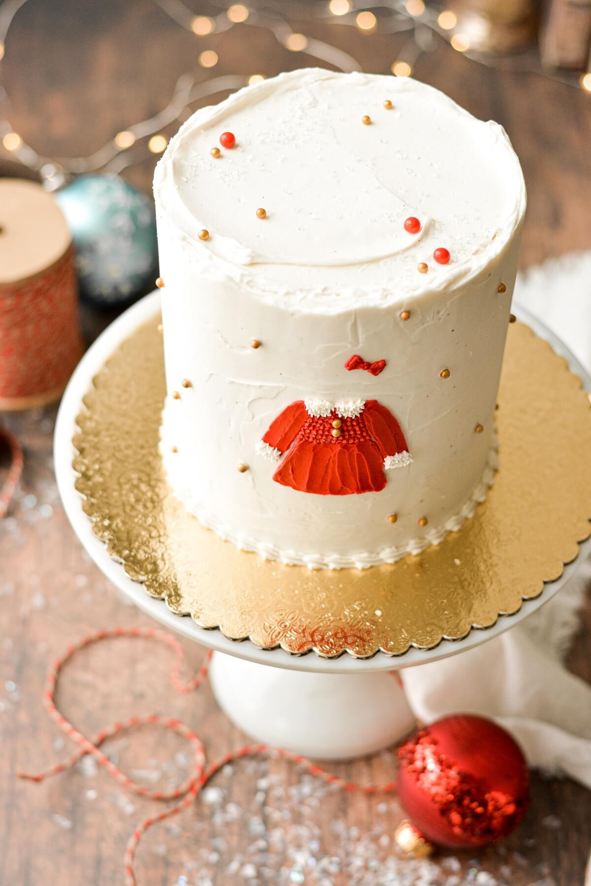 A Christmas cake with gold and red sugar pearls, ornaments, and a red Christmas dress painted on the cake in buttercream.