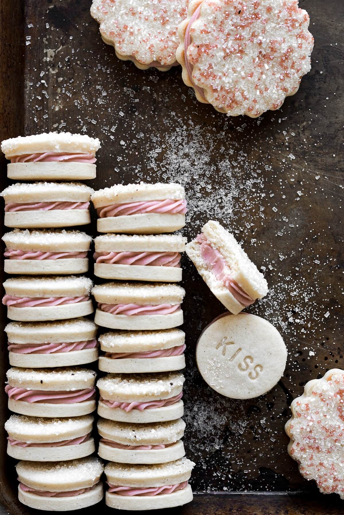 A row of vanilla bean sandwich cookies with pink raspberry frosting, on a baking sheet.