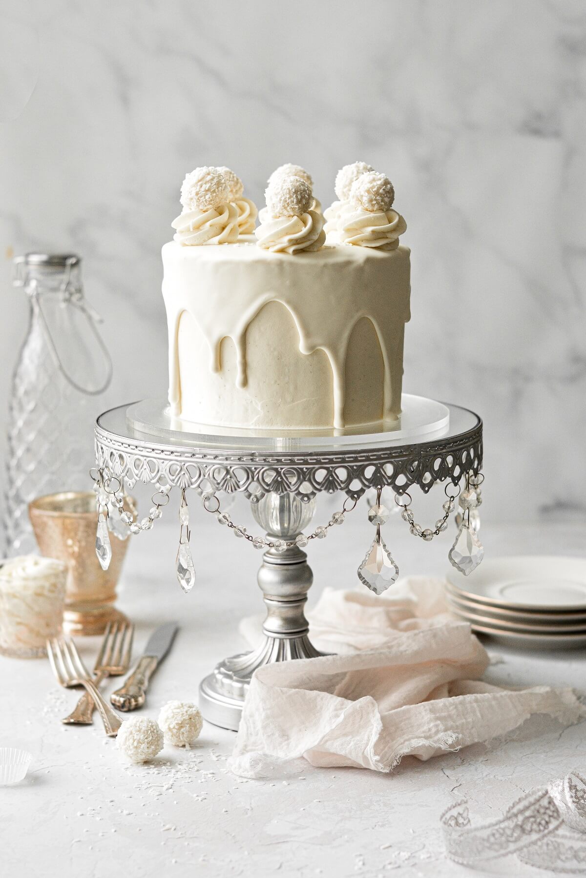 A white chocolate cake with drip and white chocolate truffles, on a silver mirrored cake stand.