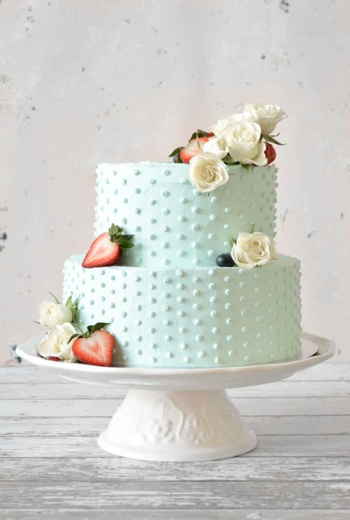 A red, white and blue, two-tiered cake with blue buttercream, swiss dots, strawberries and white roses.