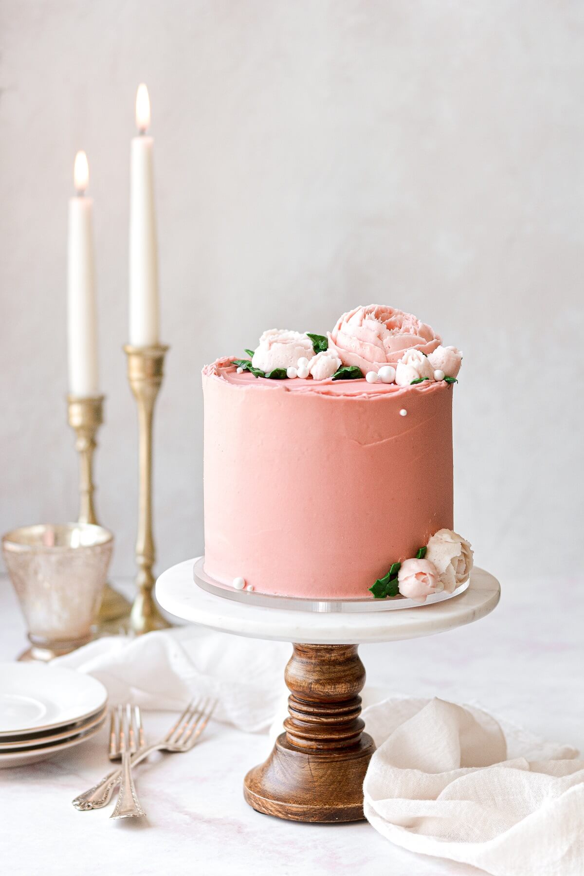 A pink frosted cake with buttercream flowers, on a wood and marble cake stand.