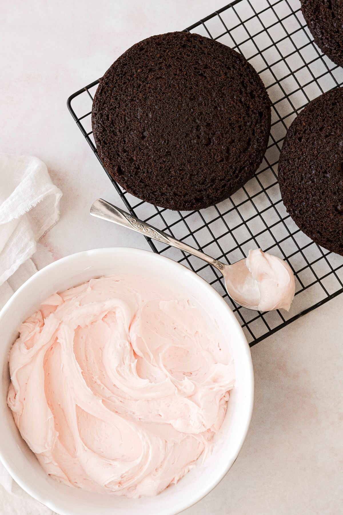 A cooling rack with layers of chocolate cake, next to a bowl of pale pink buttercream.