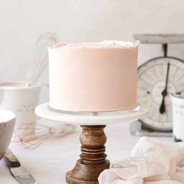 A pale pink frosted cake on a marble and wood cake stand.