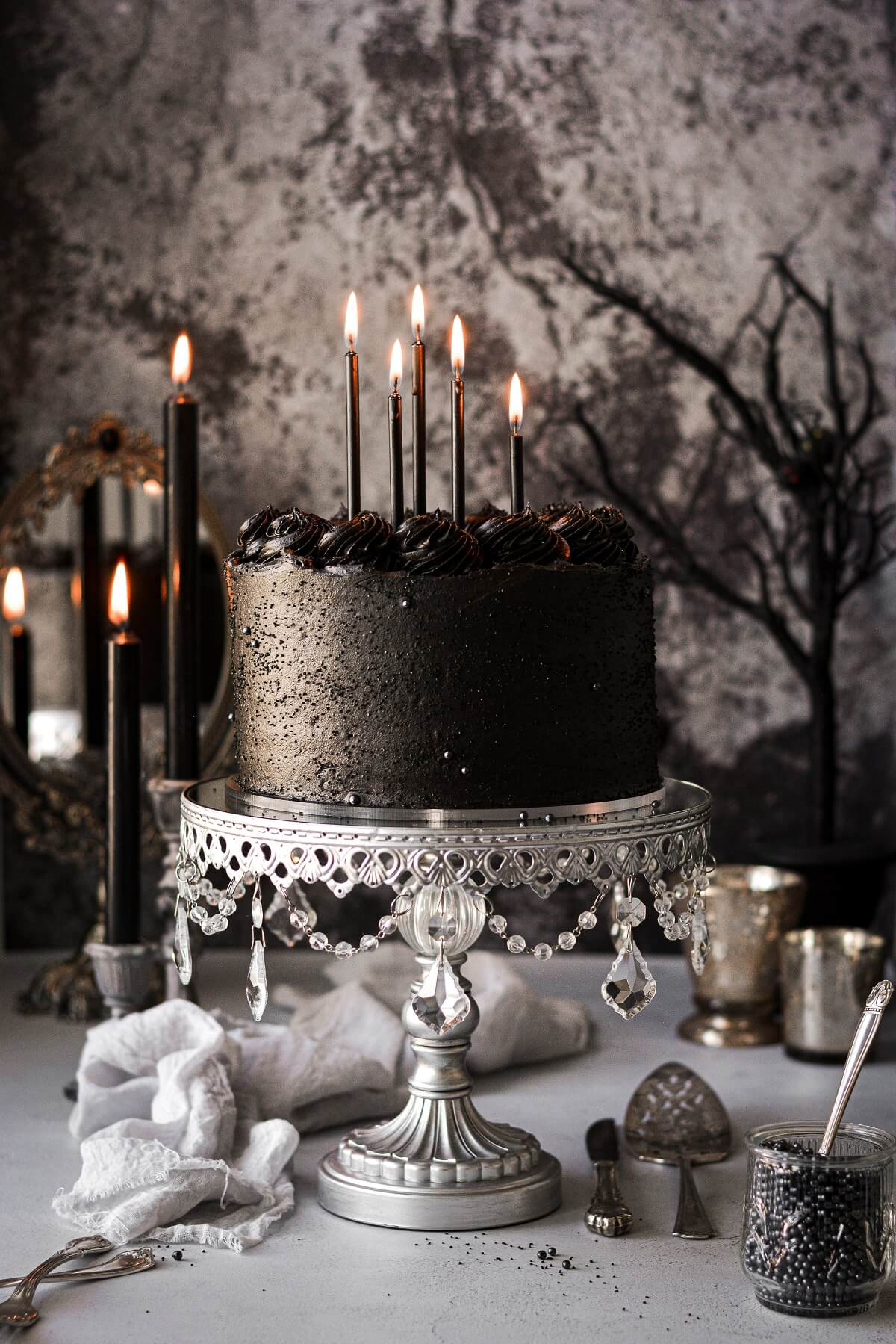 A moody scene with candles and a black velvet cake in a silver cake stand.