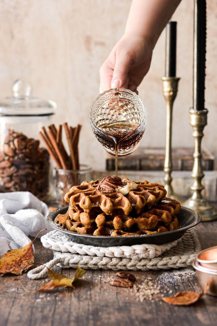 Maple syrup pouring from a glass pitcher onto a stack of pumpkin pecan waffles.
