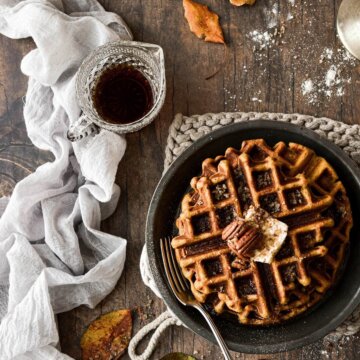 Pumpkin pecan waffles with a pitcher of syrup and leaves scattered around.