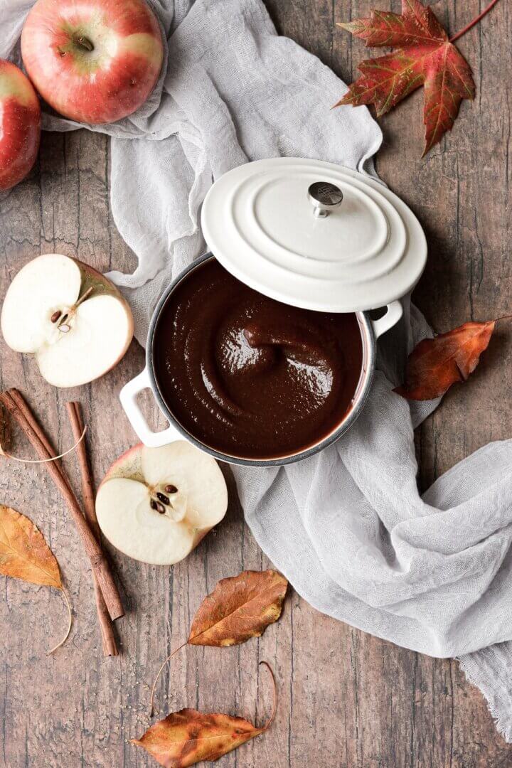 Apple butter in a white Dutch oven, surrounded by apples and leaves.