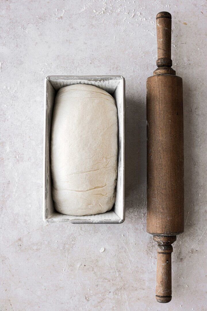 Bread dough rising in a loaf pan, next to a wooden rolling pin.