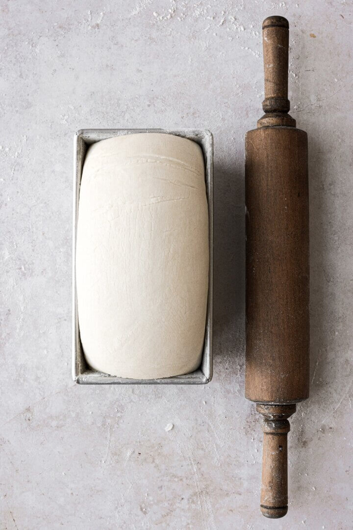 Bread dough rising in a loaf pan, next to a wooden rolling pin.