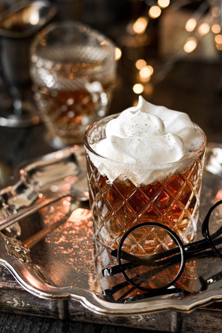 A glass of butterbeer topped with whipped cream, with lights twinkling in the background.