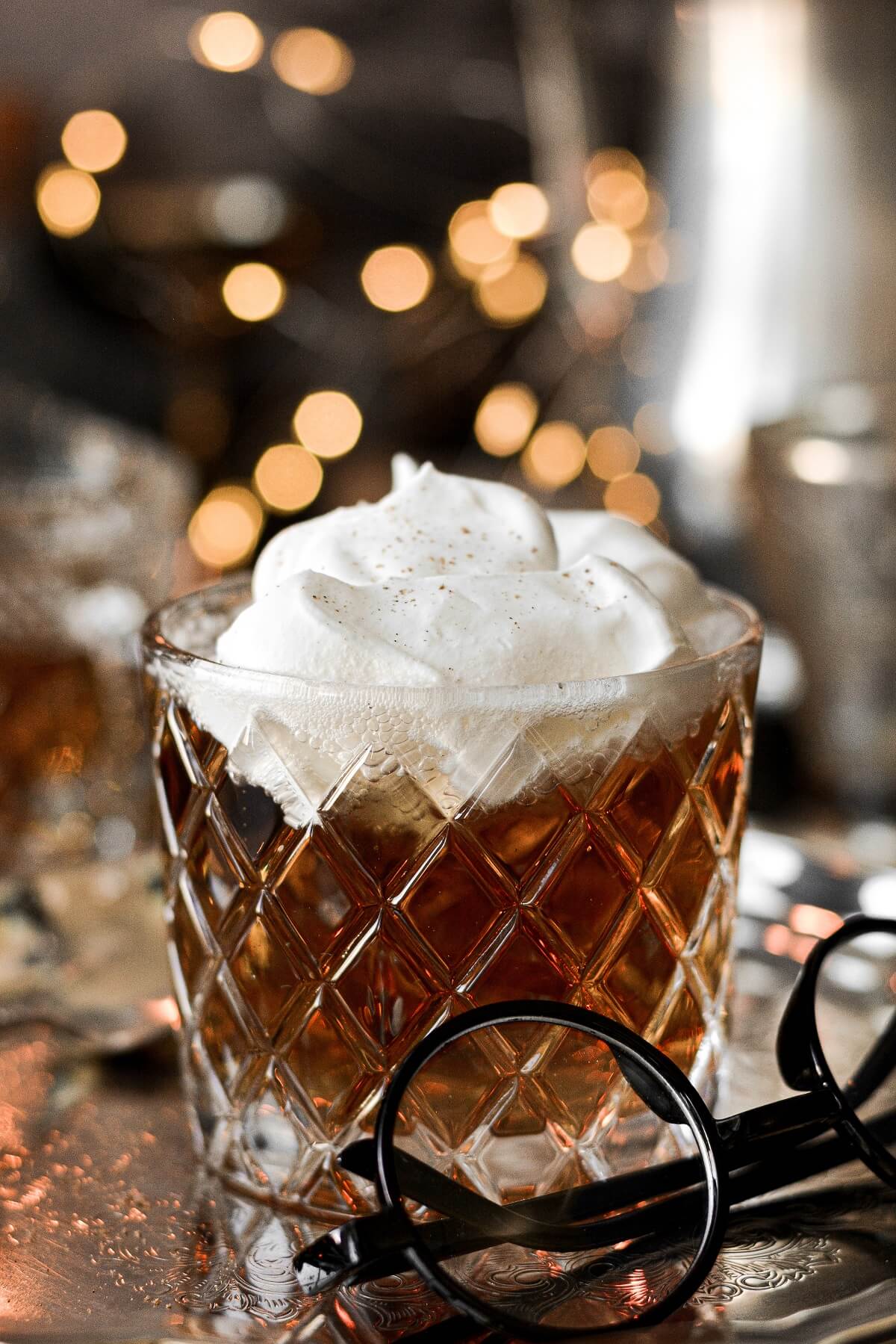 A glass of butter beer topped with whipped cream, with lights twinkling in the background.