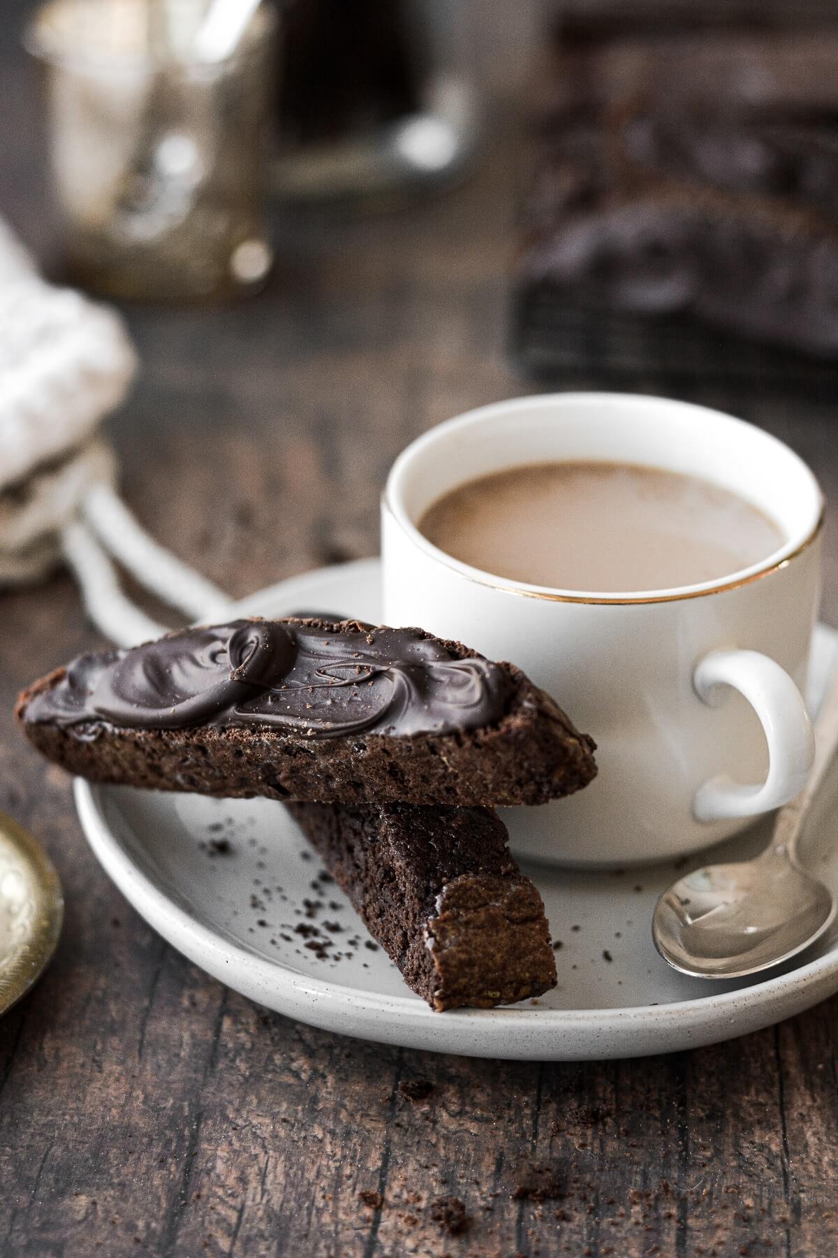 Two chocolate biscotti on a plate with a cup of coffee.