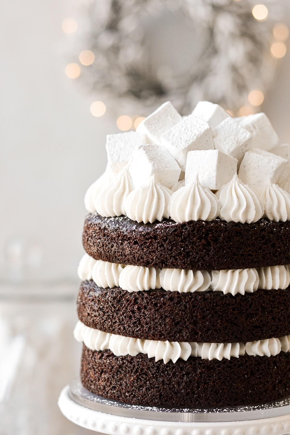 A hot chocolate cake with naked layers of dark chocolate cake, piped buttercream, and homemade marshmallows, with a lighted Christmas wreath in the background.