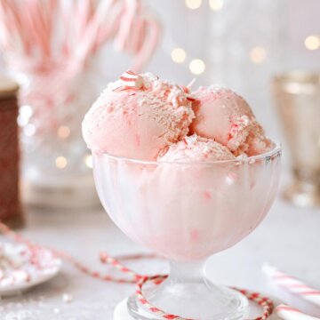 A glass footed bowl with scoops of pink peppermint ice cream, with candy canes, red twine and sparkling Christmas lights in the background.