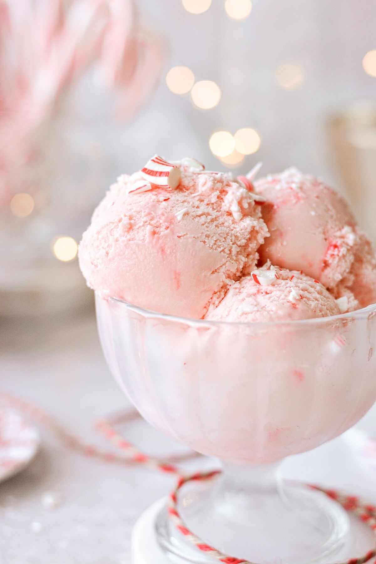 Scoops of pink peppermint ice cream in a glass footed bowl with twinkling lights in the background.
