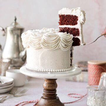 A red velvet cake on a marble and wood cake stand with a slice being lifted up.