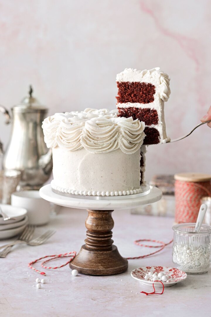 A red velvet cake on a marble and wood cake stand with a slice being lifted up.