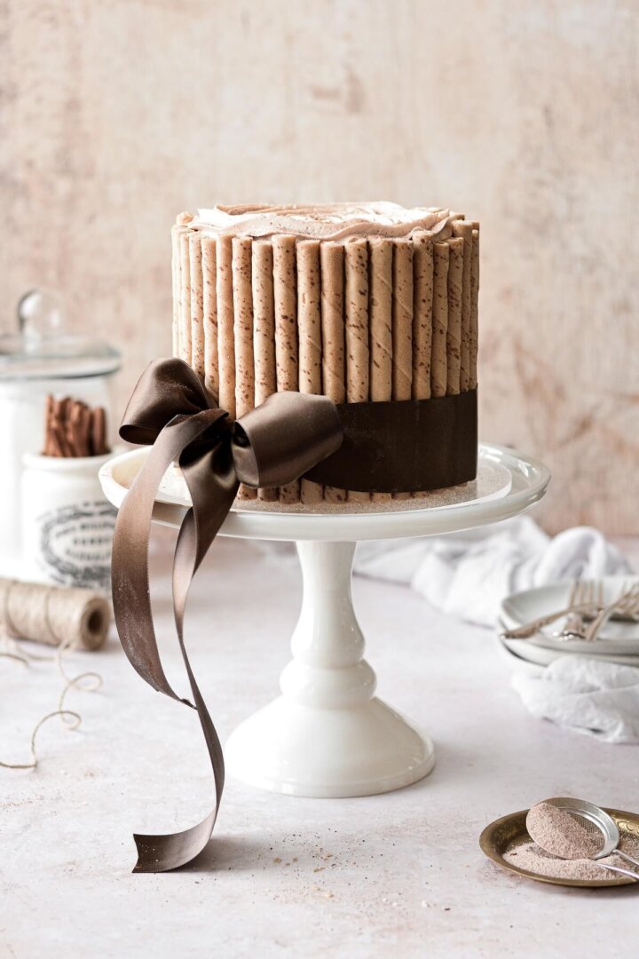 A snickerdoodle cake with piped buttercream, surrounded by Pirouettes cookies and tied with a brown satin ribbon.