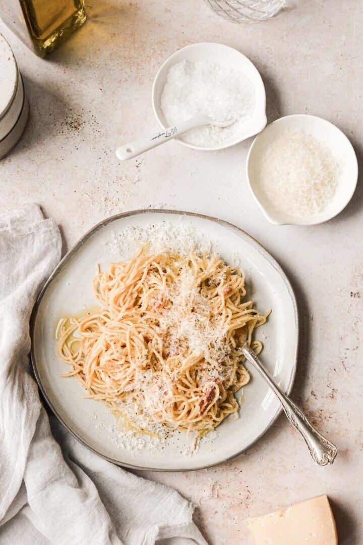 A plate of spaghetti carbonara next to bowls of salt and Parmesan cheese.