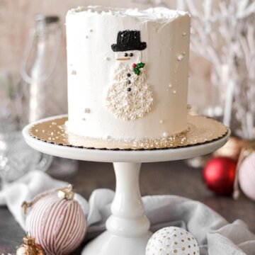 A snowman cake on a white cake stand surrounded by Christmas ornaments.