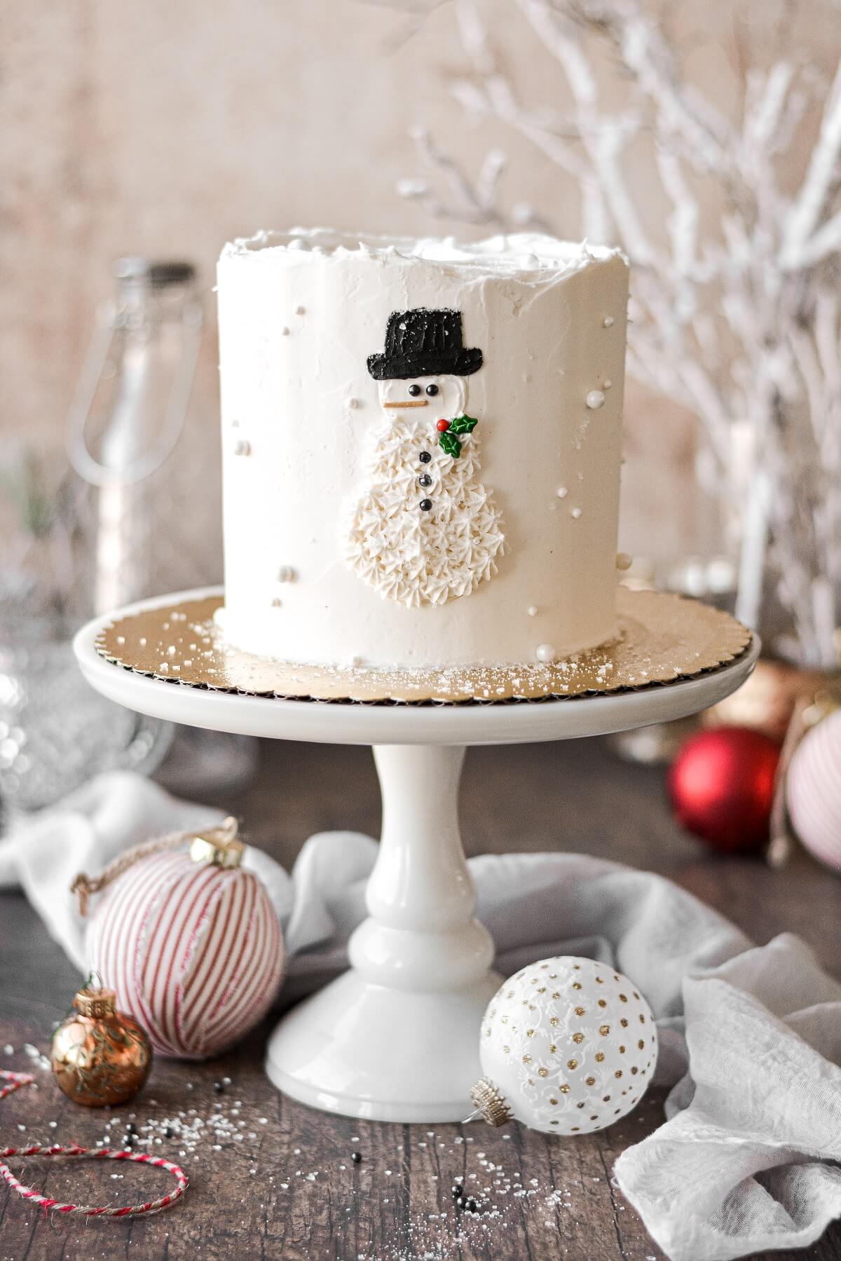 A snowman cake on a white cake stand surrounded by Christmas ornaments.