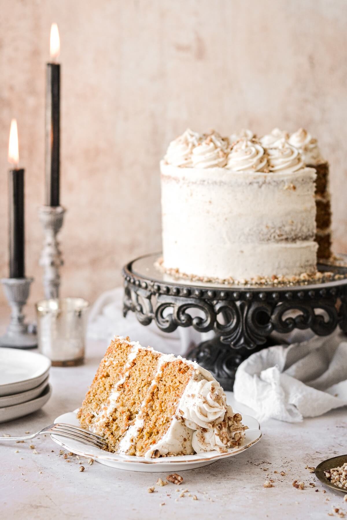 A slice of carrot cake next to a black cake stand and candles.