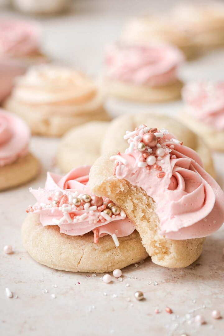 Soft sugar cookies with frosting and sprinkles, one with a bite taken.
