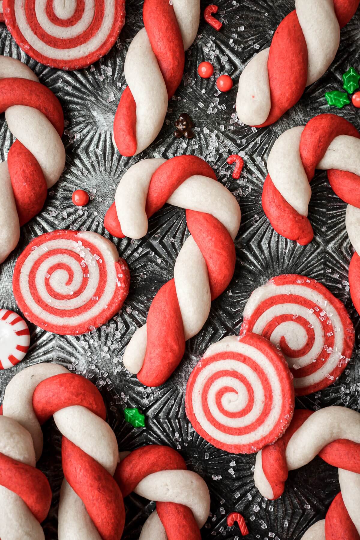 Red and white candy cane cookies and pinwheel cookies on a baking sheet with scattered Christmas sprinkles.