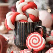 Red and white pinwheel cookies leaning against a coffee mug filled with candy cane cookies.