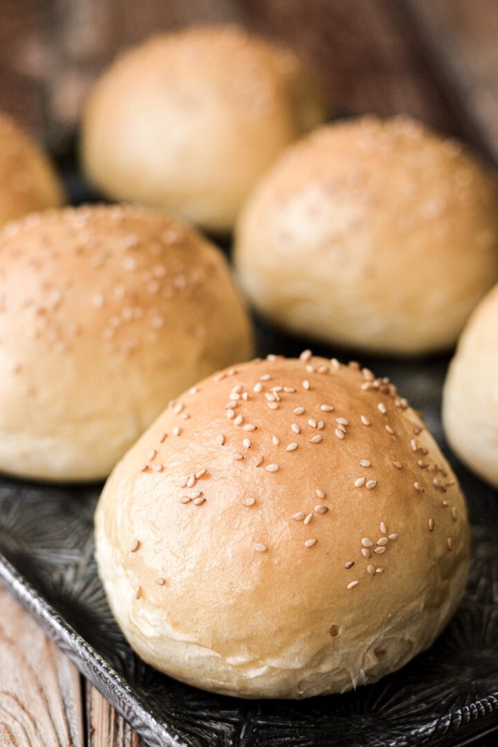 Brioche burger buns sprinkled with sesame seeds on a baking sheet.