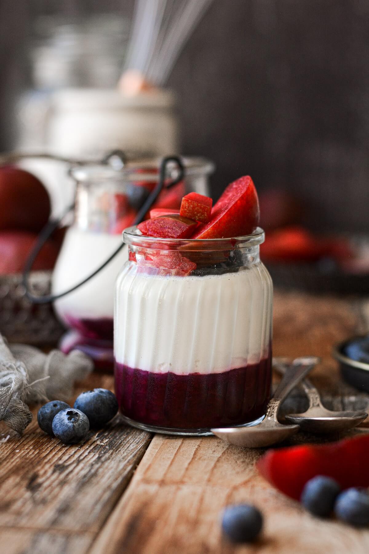 Glass jar filled with layers of fruit compote, white chocolate vanilla bean panna cotta and fresh plums and blueberries.