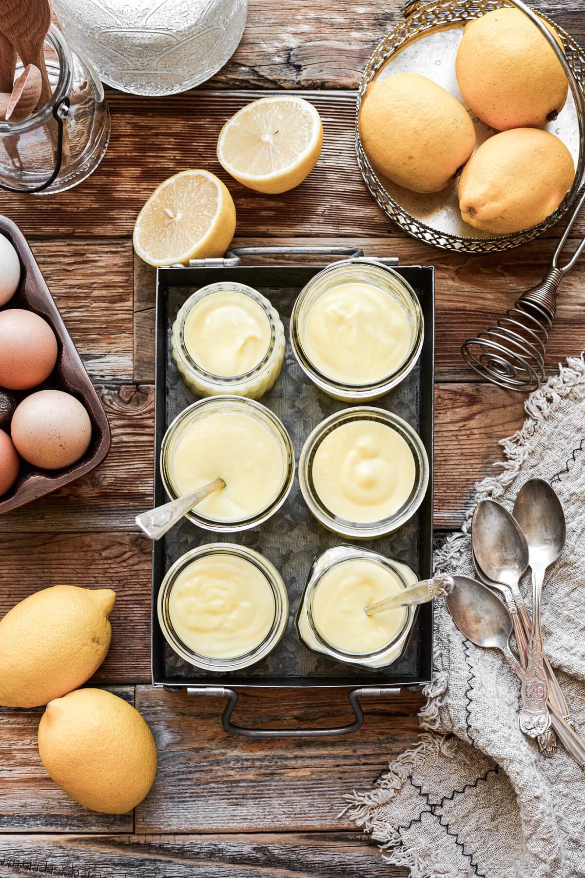 Glass jars of lemon pudding in a metal tray, surrounded by lemons.