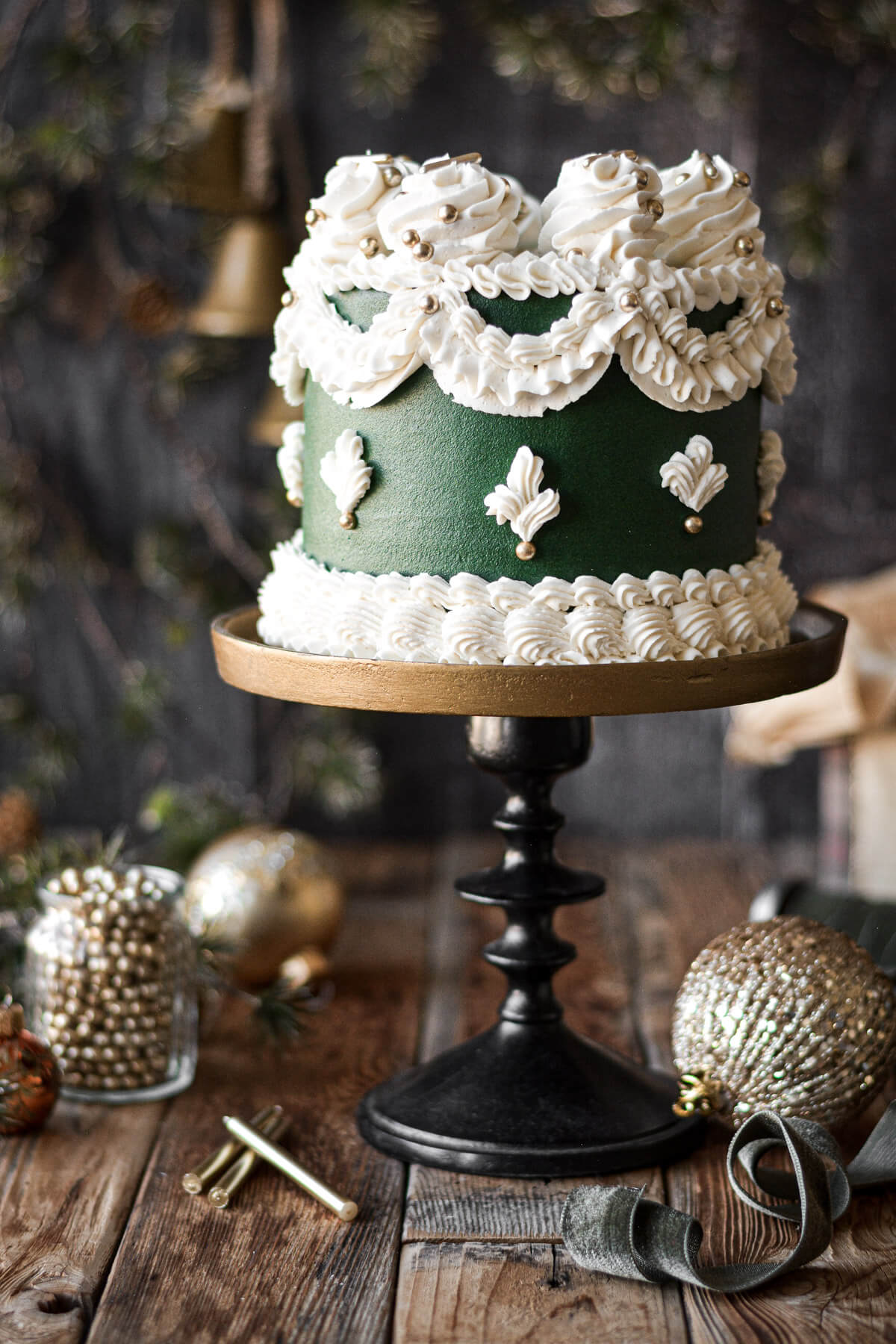 A green frosted Christmas cake with elaborate white piping, on a bronze and black cake stand.