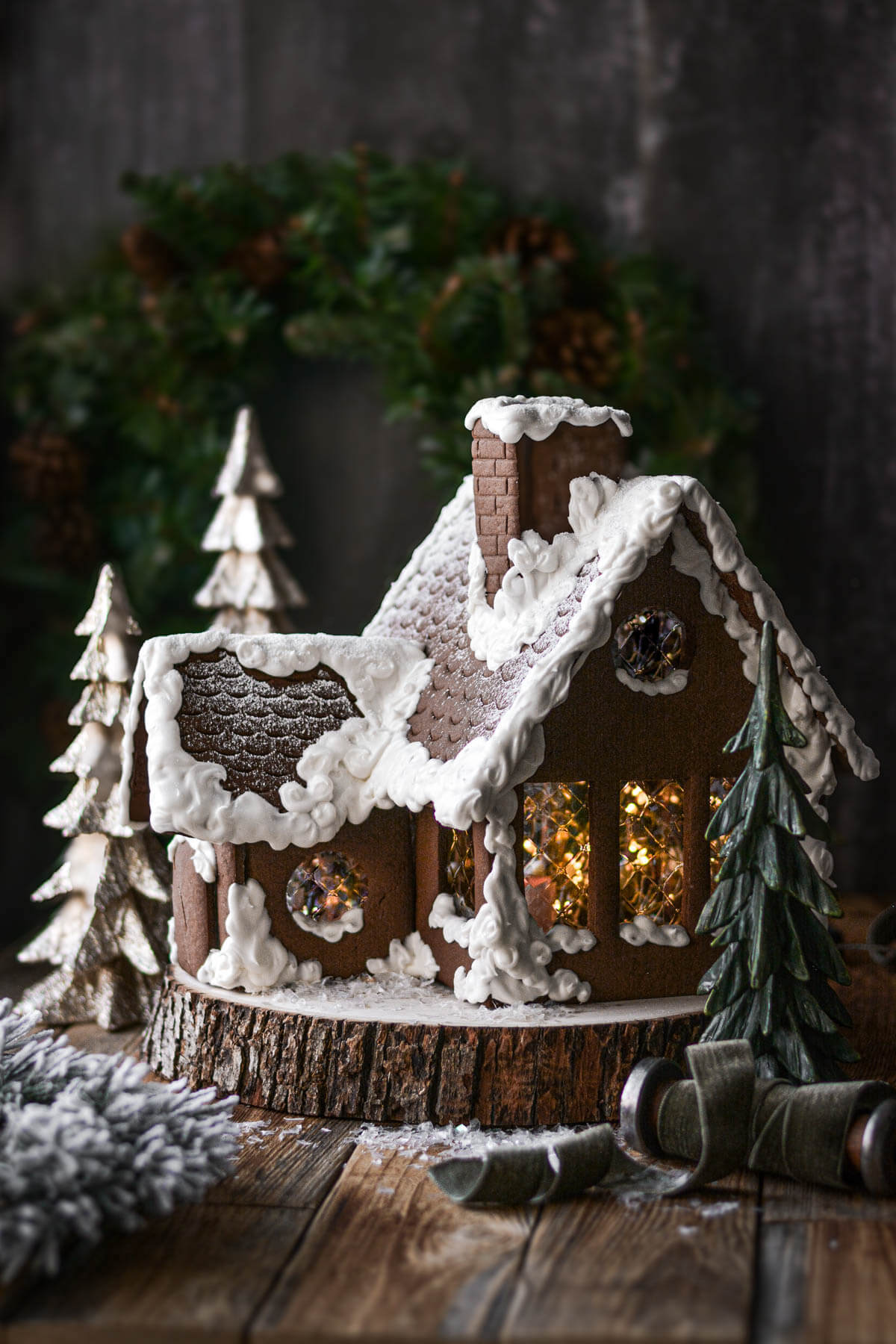 A homemade gingerbread house with icing snow, sitting on a wood slice.