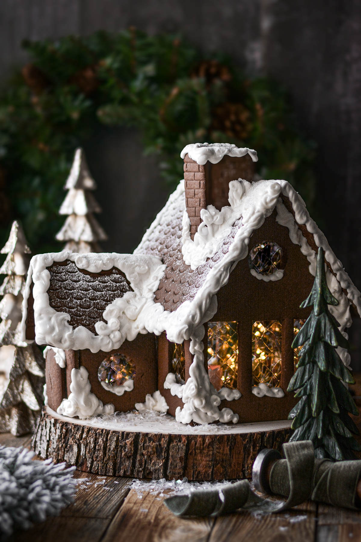 A homemade gingerbread house with icing snow, sitting on a wood slice, with lights inside the windows.