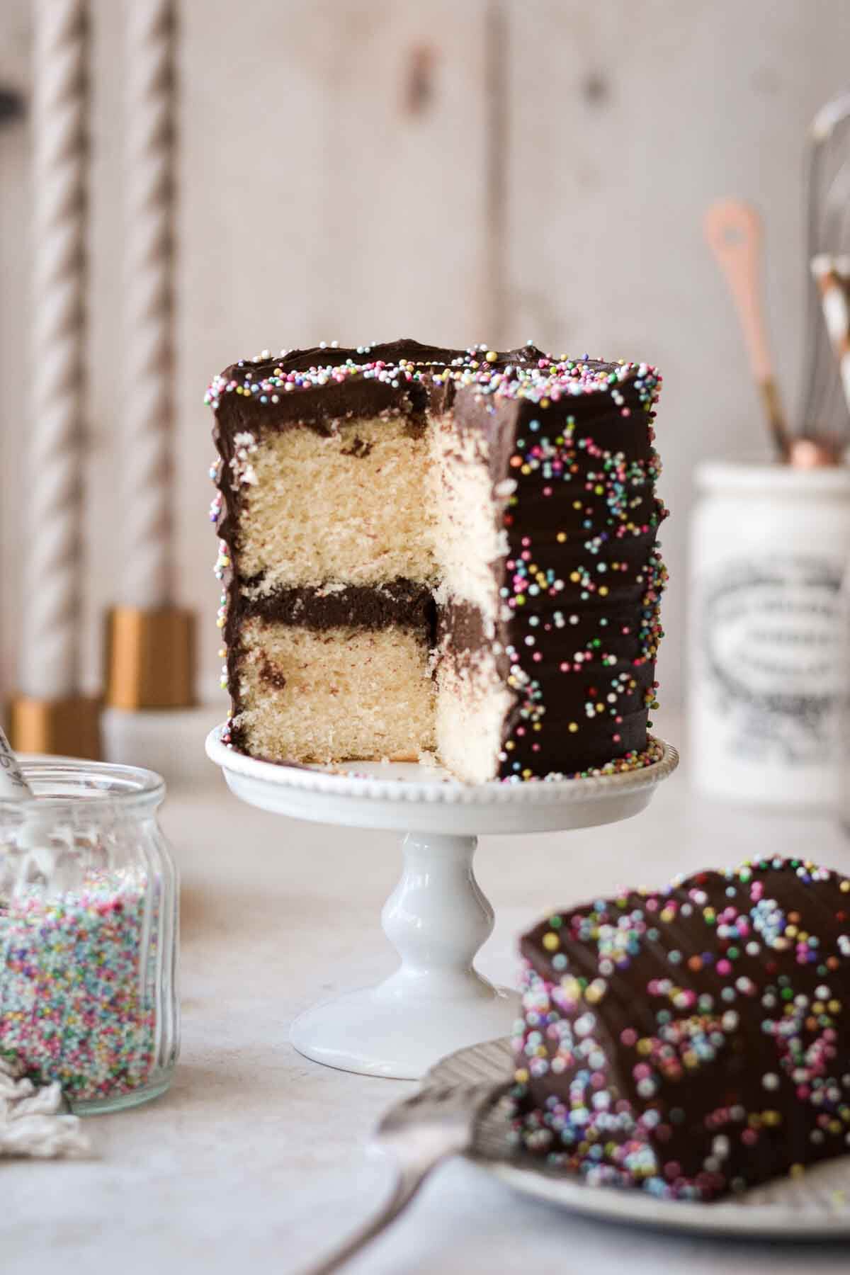 A mini yellow cake with chocolate frosting and sprinkles, with one slice cut.